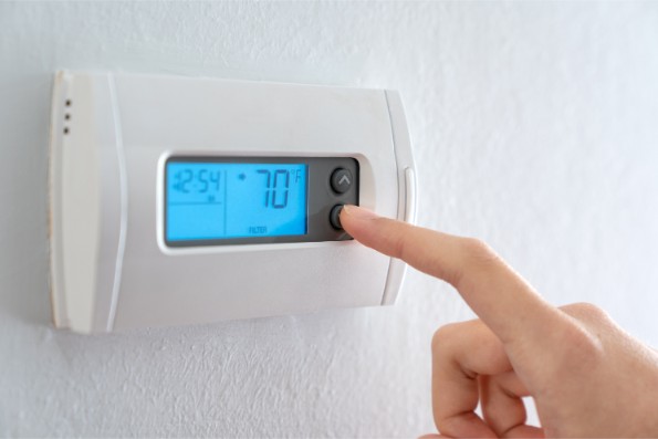 A hand adjusting the temperature on a digital thermostat mounted on a wall. The display shows a temperature of 70 degrees Fahrenheit.