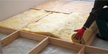 A person wearing red gloves is installing fiberglass insulation between wooden floor joists. A piece of plywood lies partially covering the insulation, and the surrounding area appears to be part of a construction or renovation project.