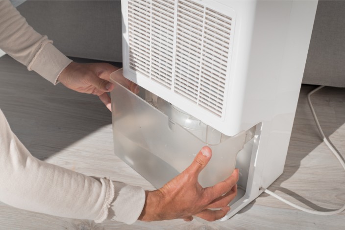 A person empties a transparent water tank from a sleek, white whole-house dehumidifier onto a light-colored wooden floor in Dallas. The unit features a front grille and is conveniently placed near the sofa, with its power cord neatly visible on the floor.