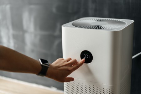 A person wearing a smartwatch adjusts a white air purifier with a circular digital display showing "0:01." The device is positioned in front of a dark, blurred background.